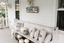 a white farmhouse porch styled for the fall, with a whitewashed bench, printed pillows, pumpkins and vintage sconces on the wall