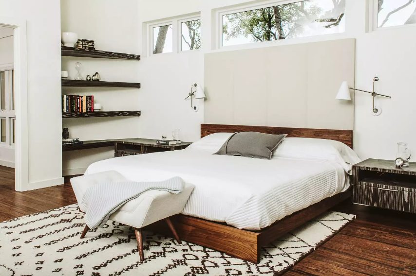 a white mid century modern bedroom with dark built in shelves, a rich stained bed, a white seat, nightstands and narrow and long windows