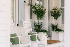 a white rustic summer porch with white furniture and planters with blooms and greenery, green pillows and a side table is very airy