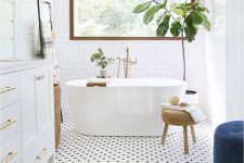 an airy mid-century modern bathroom with a penny tile floor, a white vanity and a white tub, a printed rug and a blue ottoman