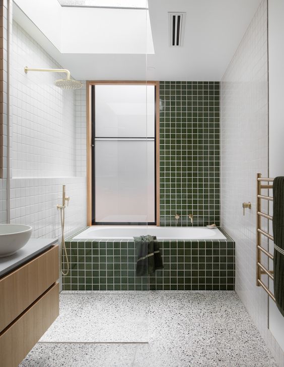 An airy mid century modern bathroom with a terrazzo floor, green and white tiles, a bathtub, a floating vanity and gold fixtures