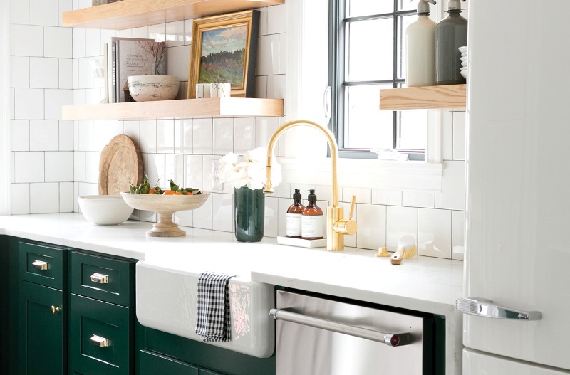 an elegant kitchen with dark green cabinets, chrome appliances, a gold faucet, gold fixtures and open thick shelves and white tiles