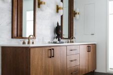 an elegant mid-century modern bathroom with marble and tan tiles, a stained wooden vanity, mirrors in wooden frames and sconces
