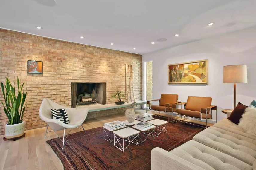 an elegant mid century modern living room with a brick wall with a fireplace, rust colored chairs, a white chair and a sofa plus an arrangement of coffee tables