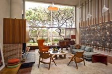 an eye-catchy living room with a fireplace clad with concrete, brick and wood, with a pale blue sofa, an orange chair, wooden chairs and a round table plus a bold artwork