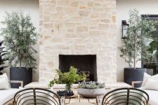 an inviting modern terrace with a large stone clad fireplace, rattan and wood furniture, potted greenery and trees is chic