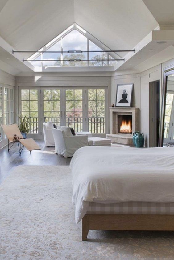 large vintage bedroom with a skylight, a fireplace and a siting zone by it, a bed with neutral bedding, an artwork and a lounger