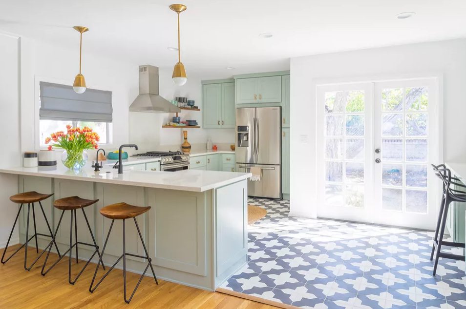 a pretty mint green kitchen with a Moroccan tile floor, brass pendant lamps, woodne stools and black fixtures is a beautiful space