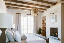 a French farmhouse bedroom with wooden beams, a built-in fireplace, built-in shelves, a neutral bed and bedding, a polished gold lamp