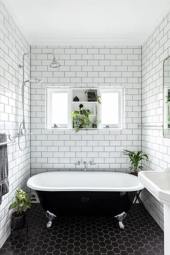 a beautiful black and white bathroom with white subway and black hex tiles, a free standing sink, a black vintage bathtub and a window