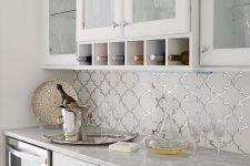 a beautiful white kitchen with glass and usual cabinets, white marble countertops, arabesque tiles accented with silver grout is wow