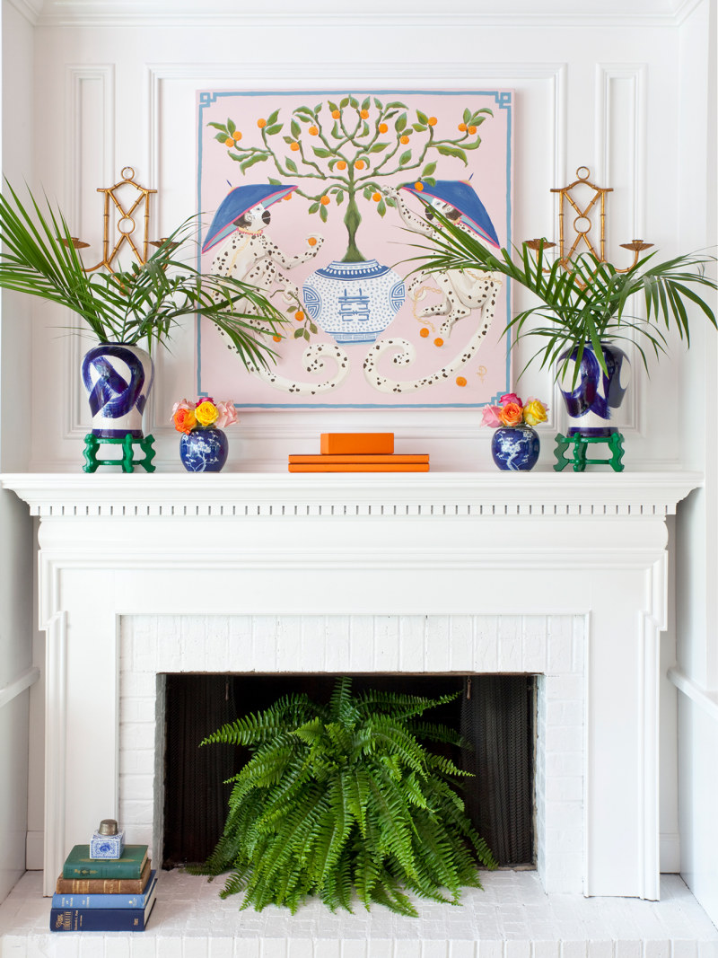 a bright space with a white fireplace and mantel, a colorful artwork, vases, blooms and greenery, a stack of books and a potted plant in the fireplace