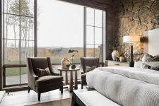 a cabin bedroom with stone walls, a bed, a dark stained bench, taupe chairs and printed pillows is welcoming