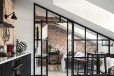 a catchy black attic kitchen with white countertops, an exposed brick backsplash, a wooden beams and a glass wall and doors leading to the bedroom