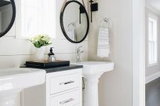 a chic farmhouse bathroom with black hex tiles, a wood plank wall, two pedestal sinks, a small storage cabinet and mirrors in black frames