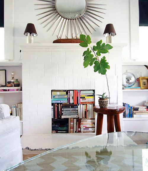 a chic living room with a white tile fireplace, books stored inside it and on the built-in shelves around, potted plants and lamps