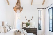 a chic neutral bedroom with wooden beams, a tan upholstered bed, neutral bedding, a crystal chandelier, a dark-stained dressser and a veiw of the lake