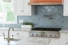 a chic vintage kitchen with white shaker cabients, a copper hood, white countertops and a blue arabesque and subway tiles on the backsplash