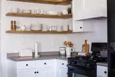 a clean white kitchen with shaker cabinets, black fixtures, grey countertops, light-stained corner floating shelves