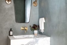 a contemporary bathroom with concrete walls, a floating stone sink, a catchy mirror, gold fixtures and brass sconces is very chic