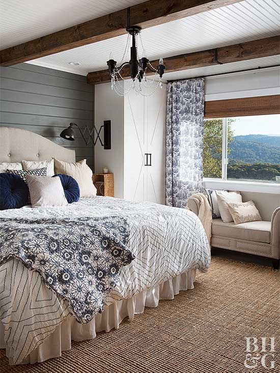 a cool farmhouse bedroom with rich-stained wooden beams, a neutral upholstered bed, a neutral sofa, printed bedding and a white wardrobe