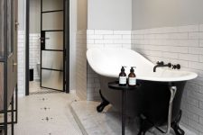 a cool modern bathroom with white subway tiles and penny ones, a black clawfoot tub, a brown vanty and a side table is all chic