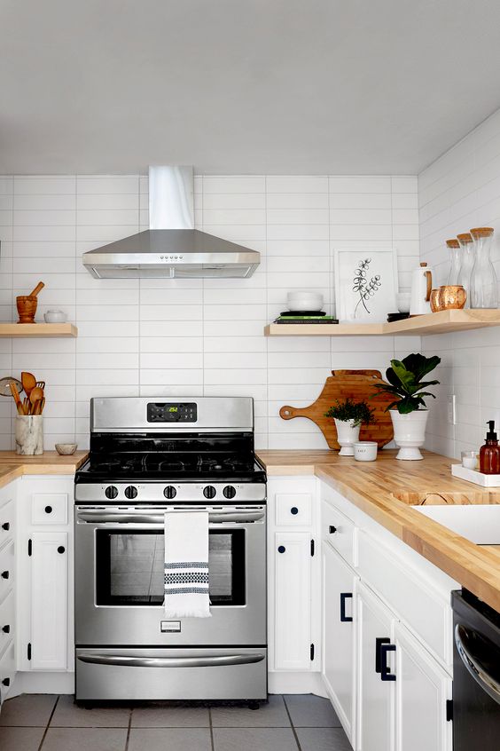 a cute white kitchen with black fixtures, light stained butcherblock countertops and floating shelves plus a skinny tile backsplash