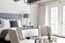 a farmhouse bedroom in neutrals,w ith rich-stained wooden beams, a metal sphere chandelier, a grey bed and creamy chairs, a printed rug