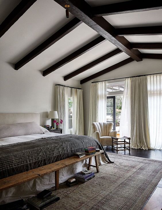 a farmhouse bedroom with dark wooden beams, a neutral bed and a wooden bench, a creamy char, neutral bedidng and a printed rug