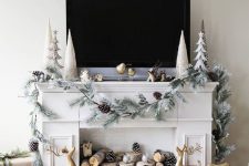 a faux fireplace styled for Christmas, with firewood, pinecones, gold deer, a whitewashed evergreen garland with pinecones and pretty trees