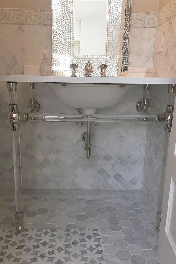 a glam bathroom clad with marble arabesque and hexagon tiles, with an acrylic sink stand and a mirror in a shiny wide frame is cool