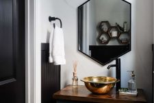 a glam powder room with a dark green vanity, a gold vessel sink, a hexagon mirror in a black frame, neutral nextiles and a large sconce