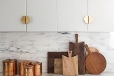 a grey and white kitchen with elegant gold knobs, with white marble countertops and a backsplash, with copper jars is a chic idea