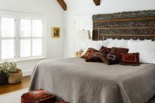 a historic farmhouse bedroom with rich-stained wooden beams, a bed with a carved headboard, neutral bedding and bold pillows, bold printed stools