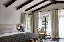 a large and cool neutral bedroom with dark wooden beams on the ceiling, a neutral bed, a wodoen bench and neutral textiles