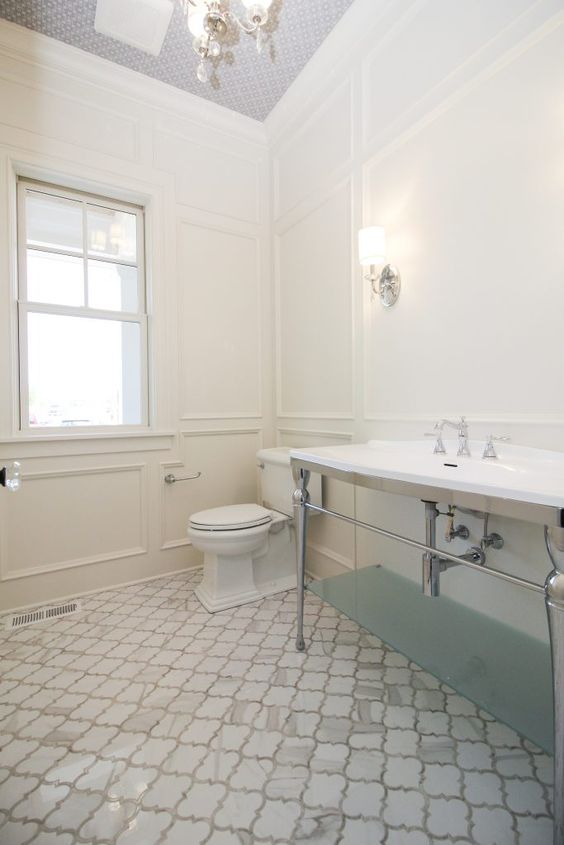 a large neutral vintage bathroom with paneled walls, white marble arabesque tiles on the floor, a large sink stand and a printed ceiling