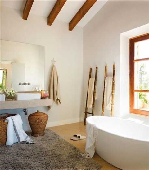 a lovely coastal bathroom with rich-stained wooden beams and a window frame, an oval tub, a floating vanity, baskets for storage and ladders