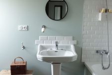 a lovely vintage bathroom with green walls, a bathtub with subway tiles around, a vintage pedestal sink with subway tiles, a round mirror, a mini stool and a chest for storage
