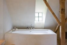 a minimalist neutral bathroom with a wooden pillar and a wooden beam, a bathtub clad with white stone, a wooden floor and a small window