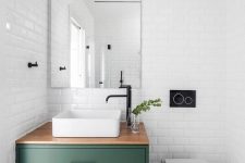 a modern and serene powder room clad with white subway tiles, with a green and black vanity, a square vessel sink, white appliances and a large mirror