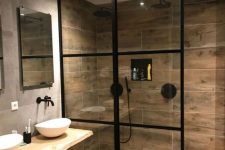 a modern bathroom clad with grey and wood look tiles, with black fixtures, bowl sinks, a framed shower wall is a cool and cozy space