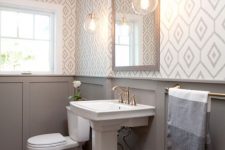 a modern farmhouse bathroom with geo wallpaper, grey paneling, a pedestal sink, a mirror in a wooden frame and pendant lamps