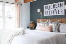 a modern farmhouse bedroom with rich-stained wooden beams on the ceiling, a stained bed, neutral bedding, a black accent wall and a printed rug