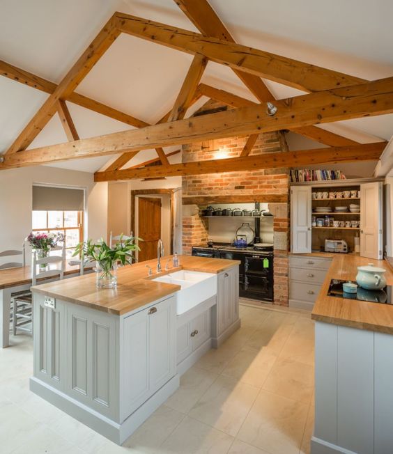 a modern farmhouse kitchen with grey furniture, light-stained butcherblock countertops, an exposed brick wall and wooden beams