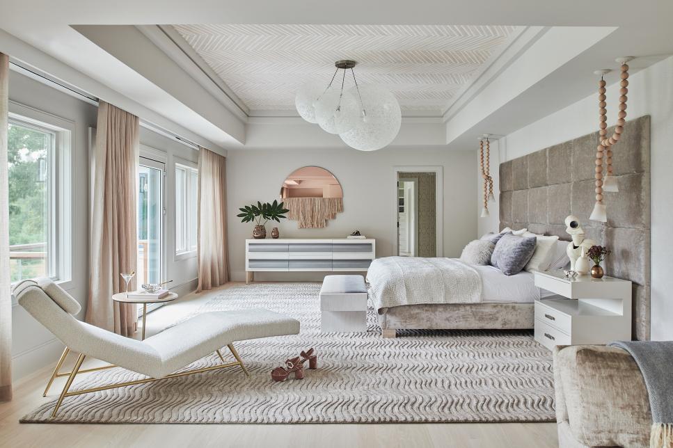 a modern refined bedroom in neutrals, with anupholstered bed, neutral bedding, a cool dresser, a creamy lounger, blush curtains and a copper mirror
