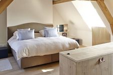 a modern rustic attci bedroom with wooden beams, a neutral upholstered bed, neutral bedding and nneutral planked furniture