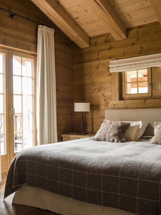 a modern rustic bedroom clad with wood, with wooden beams, an upholstered bed with neutral bedding, wooden nightstands with lamps
