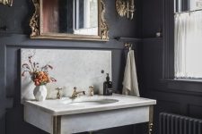 a moody powder room with black walls and paneling, a console sink, a mirror in an ornated frame, sconces and neutral textiles