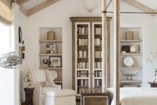 a neutral French farmhouse bedroom with wooden beams, a stained bookcase, built-in shelves, a cnaopy bed and a neutral chair
