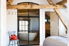 a neutral bedroom with light-stained wooden beams, a bed with neutral bedding, a pendant lamp and a grey chair plus a window to the bathroom